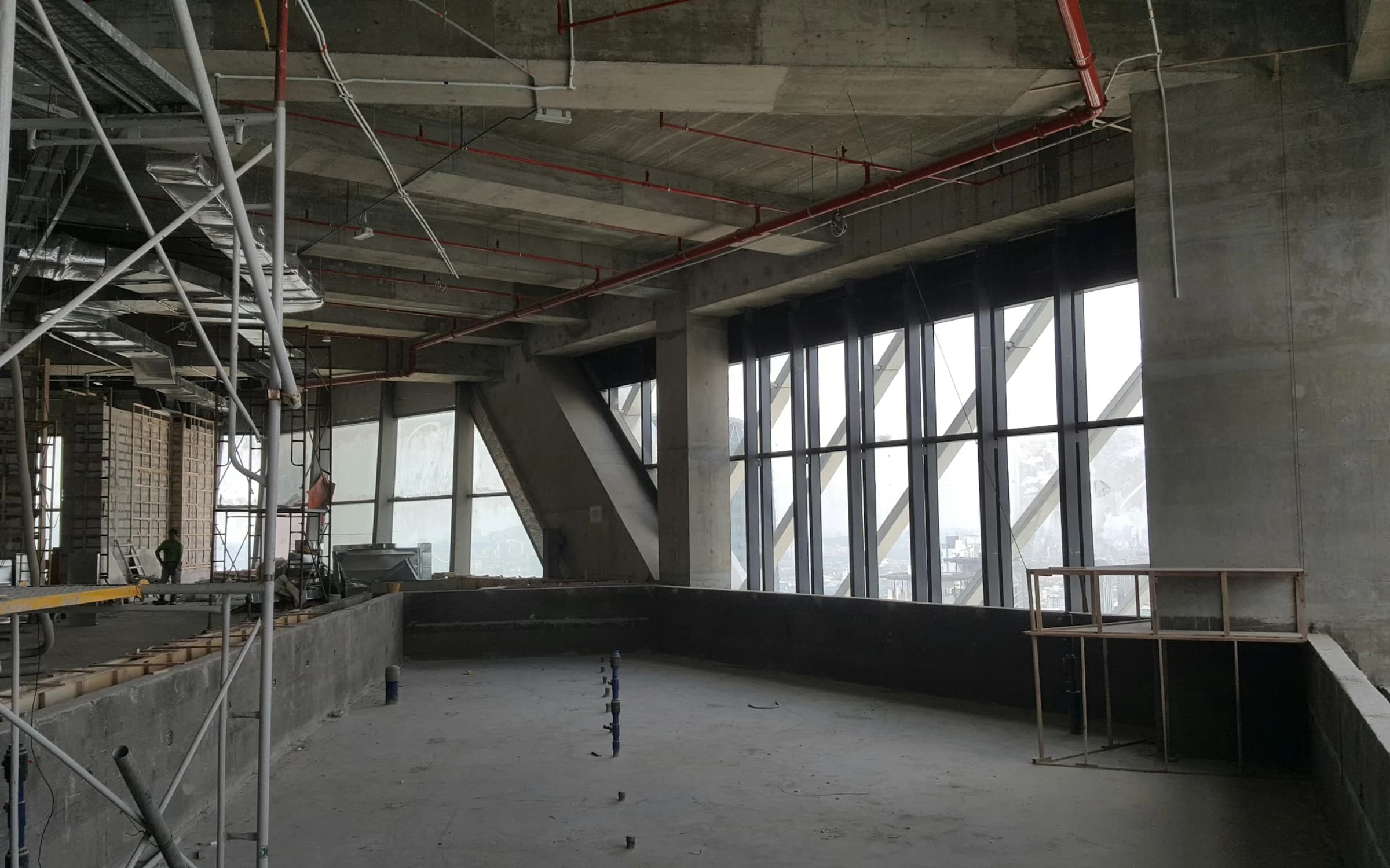 Interior de concreto en obra con andamios, instalaciones expuestas y grandes ventanales.